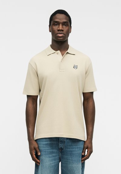 Homme portant un polo beige à manches courtes avec un petit logo de loup sur la poitrine et un jean bleu, debout devant un fond uni.