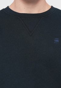 Felpa blu navy con collo a girocollo a coste, dettaglio a V sul colletto e piccolo logo sul petto. Realizzata in tessuto morbido e testurizzato.