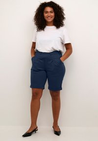Shorts blu navy su misura con risvolti, pieghe davanti e tasche laterali, abbinati a una maglietta bianca semplice e tacchi neri.