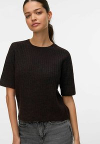 Vrouw met donker paardenstaartje, gekleed in een zwart geribd kortemouwtruitje en grijze spijkerbroek, tegen een effen witte achtergrond.