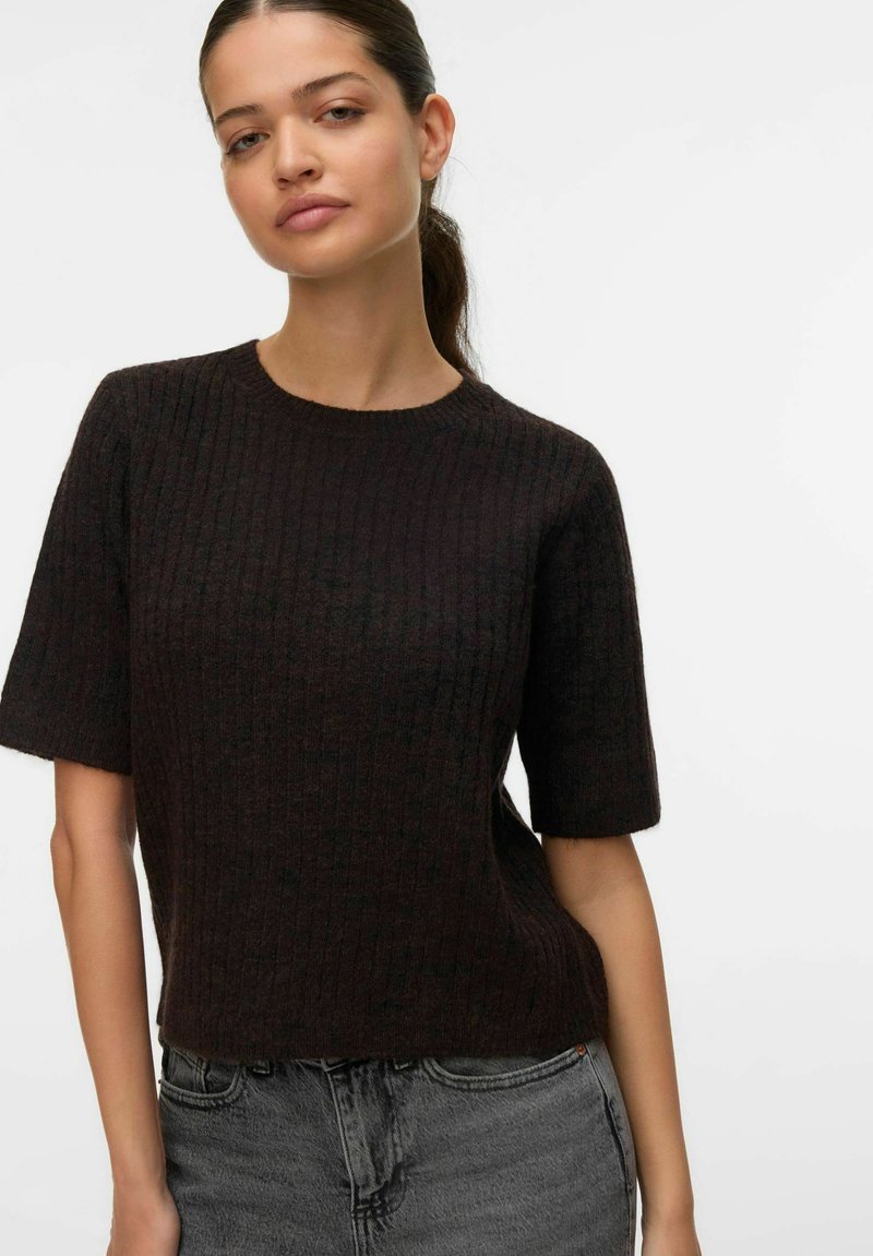 Vrouw met donker paardenstaartje, gekleed in een zwart geribd kortemouwtruitje en grijze spijkerbroek, tegen een effen witte achtergrond.