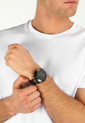 Uomo con maglietta bianca che indossa un orologio da polso nero in metallo, che lo sistema sul polso sinistro con la mano destra.