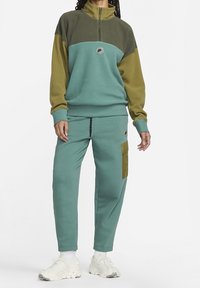 Fleece-pullover i grön och teal med en kvartdragkedja och logotyp. Matchande tealbyxor med en ficka på sidan och manschetter vid anklarna. Vita sneakers.