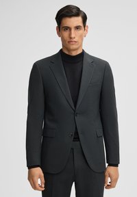 Donkergrijze op maat gemaakte colbert met notch-lapels, twee knopen en zijzakken. Gemaakt om te dragen over een zwarte coltrui, gemaakt van gladde stof.