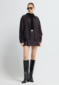 Paarse gestreepte jacket met rits, zwarte coltrui en minirok; kniehoge zwarte laarzen met krokodillenprint, geaccessoireerd met een zonnebril.