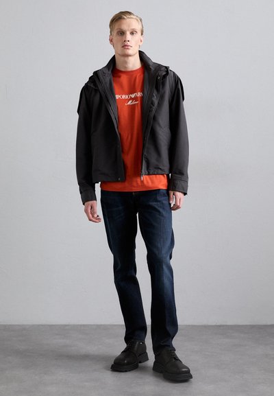 Black jacket with a hood over an orange t-shirt featuring white text, paired with dark blue jeans and black shoes. Simple design and materials.