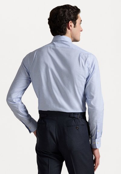 Cămașă în carouri de culoare albastru deschis, cu guler larg și manșete cu nasturi, purtată cu pantaloni negri croiți, care au curele laterale ajustabile.