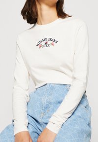 Krémová košile s dlouhým rukávem, na které je vyšitý nápis "TOMMY JEANS N.Y.C." tmavým textem a růžovými růžemi. Nositelná s světle modrými džínami s vysokým pasem a vzorem.