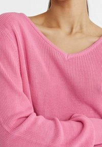 Pull rose en tricot avec un large col en V, une texture côtelée et des manches longues, mettant en valeur un matériau doux et léger.