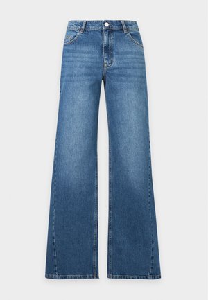 Vrouw staand met handen achter de rug, dragend een beige trui en wijde blauwe jeans tegen een effen lichte achtergrond.