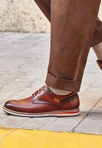 Zapato de cuero marrón con cordones naranjas, suela a rayas blanca y roja que contrasta, y punta redonda. Usado con pantalones marrones remangados y calcetines claros.