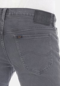 Jeans in denim grigio con un'etichetta in pelle nera sulla vita e un piccolo tag arancione. Presenta tasche posteriori classiche con cuciture curve.