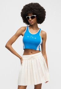Femme aux cheveux afro portant des lunettes de soleil blanches, un crop top Nike bleu et une jupe plissée blanche, se tenant avec une main sur la hanche.