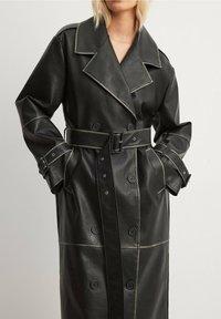 Schwarzer Leder-Trenchcoat mit doppelter Knopfleiste, kontrastierender Naht, großen Revers und tailliertem Gürtel. Verfügt über eingearbeitete Taschen mit Knöpfen.