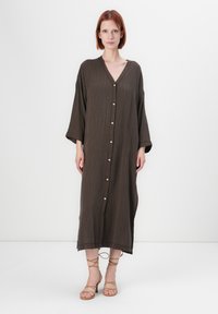 Une femme se tient face à l'avant, portant une longue robe brune, loose, à boutons avec de larges manches et des sandales à talons beiges lacées.