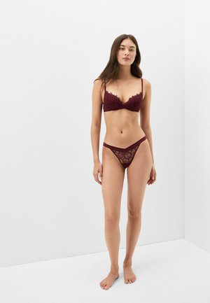 Jeune femme debout pieds nus sur un fond blanc uni, portant un soutien-gorge en dentelle bordeaux et un sous-vêtement assorti.