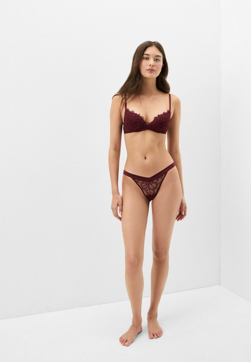 Young woman standing barefoot against a plain white background wearing burgundy lace bra and matching underwear.