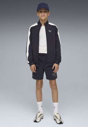 Niño con chaqueta deportiva azul marino y pantalones cortos, camiseta blanca, calcetines blancos de Puma, gorra y zapatillas de correr, de pie frente a un fondo gris.