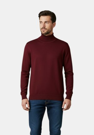 Uomo in piedi che indossa un maglione dolcevita a maniche lunghe color bordeaux e jeans blu, guarda leggermente verso sinistra.