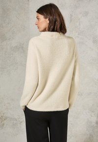 Pull en beige clair, texturé, avec une coupe ample et un col montant ; associé à un pantalon noir, montrant un matériau doux et confortable.