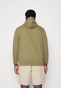 Felpa verde oliva con tasca frontale e cappuccio grande, abbinata a pantaloncini beige. Il tessuto sembra morbido, con polsini e orlo a coste.