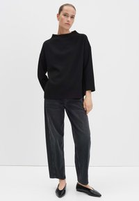 Frau in einem lockeren schwarzen Longsleeve-Oberteil, verwaschenen schwarzen geraden Jeans und schwarzen spitzen Ballerinas vor einem schlichten weißen Hintergrund.