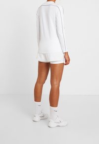 Weißes Langarmshirt mit schwarzen Paspeln, kombiniert mit weißen Shorts. Person trägt weiße Nike-Schuhe und Socken und steht vor einem einfarbigen Hintergrund.