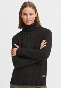 Pull à col roulé en tricot noir avec texture en maille torsadée, poignets et ourlet côtelés, et un petit patch en cuir au bas sur le côté.