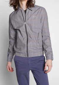 Mężczyzna ubrany w szarą, kraciastą kurtkę zapinaną na zamek z klapowymi kieszeniami oraz fioletowe spodnie, stojący na tle jednolitego jasnego tła.