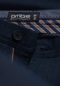 Tessuto blu navy con un motivo testurizzato; presenta un bottone nero e una cerniera dorata. L'etichetta riporta "ombre QUINTESSENCE M/32." I dettagli includono una fodera a righe.