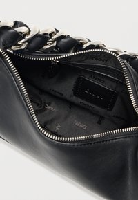 Μαύρη δερμάτινη τσάντα με ασημένια αλυσίδα, κλείσιμο με φερμουάρ και υφασμάτινη επένδυση με ανάγλυφο λογότυπο. Το εσωτερικό διαθέτει μια μικρή τσέπη με φερμουάρ.