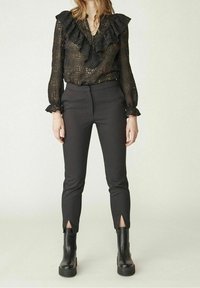 Femme portant un blouse en dentelle noire à manches longues avec volants, un pantalon noir ajusté avec des fentes au niveau des chevilles et des bottes noires épaisses, debout sur un fond uni.