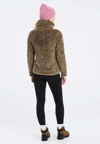 Bruine fuzzy fleece jas met een hoge kraag, zwarte legging, en een roze beanie, gecombineerd met bruine wandelschoenen. Achteraanzicht.