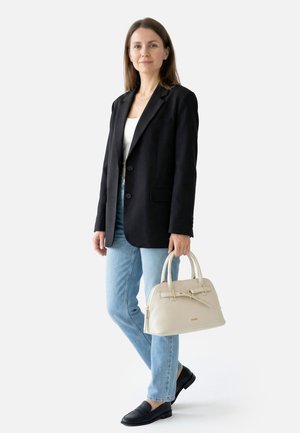 Žena stojící, oblečená v černém saku, bílém topu, světle modrých džínách a černých mokasínách, držící krémově zbarvenou kabelku se zlatými doplňky.