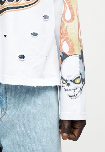 Άσπρο μακρυμάνικο πουκάμισο με τρύπες και πορτοκαλί φλόγα και σχέδιο κρανίου στο μανίκι, φορεμένο με ανοιχτόχρωμο μπλε τζιν.