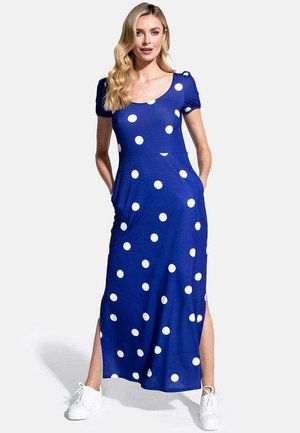 Žena v modrých maxi šatách s bielymi bodkami, krátkymi rukávmi, bočnými rázporkami a bielymi teniskami, stojaca s rukami vo vreckách.