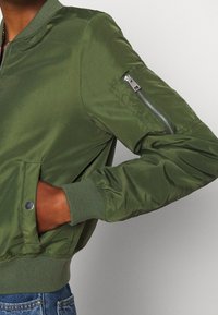 Kurtka bomberka w kolorze oliwkowej zieleni z kieszenią na zamek na lewym rękawie, ściągaczami na mankietach oraz gładką, błyszczącą fakturą tkaniny.
