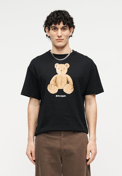 Junger Mann mit lockigem Haar, der ein schwarzes T-Shirt mit einer großen Teddybär-Grafik und braune Hosen vor einfarbigem Hintergrund trägt.