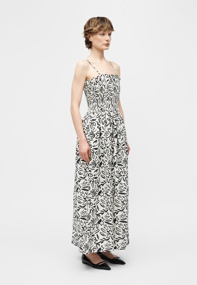 Γυναίκα που φοράει μακριά μαύρη και λευκή φλοράλ φόρεμα με λεπτές τιράντες και μαύρα μυτερά φλατ παπούτσια, στέκεται μπροστά σε απλό φόντο.