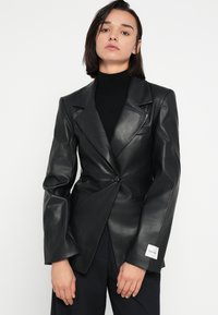 Čierny kožený blejzer s pevne tvarovanými ramenami, zapínaním na jeden gombík a prednými vreckami. Na ľavom rukáve je značka značky.