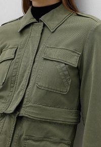Veste vert olive avec un col, fermeture à bouton et quatre poches avant. Fabriquée en tissu durable avec une finition texturée, dotée de coutures visibles.