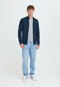 Mornarska zip-up cardigan preko sive majice, svijetloplave traperice i bijele tenisice s crnim prugama, stoji uz svijetlu pozadinu.