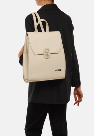 Zaino in pelle beige con un design strutturato, caratterizzato da hardware in rilievo e due manici superiori. Indossato sulla spalla abbinato a un outfit scuro.