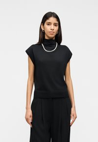 Femeie cu păr drept și închis la culoare, poartă un top negru fără mâneci cu gât înalt, pantaloni negri plisati, cercei rotunzi și un colier simplu de perle.