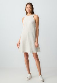 Ein ärmelloses Leinenkleid in hellem Beige, mit lockerem Schnitt und weit auslaufendem Saum, kombiniert mit weißen Turnschuhen für einen lässigen Look.