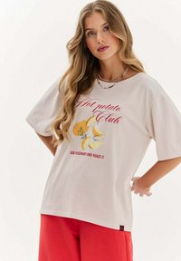 Krémové tričko s krátkým rukávem s grafikou žlutých brambor a bylinek, text "Hot Potato Club" v červené barvě. Měkká bavlněná tkanina.