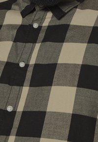 Camisa de cuadros abotonada en negro y beige. Presenta un cuello clásico, bajo redondeado y botones blancos, con una tela texturizada.