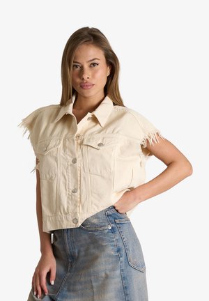 Giovane donna che posa con la mano sui fianchi, indossando una giacca di jeans beige senza maniche con bordi sfrangiati e una gonna di jeans blu sbiadita.