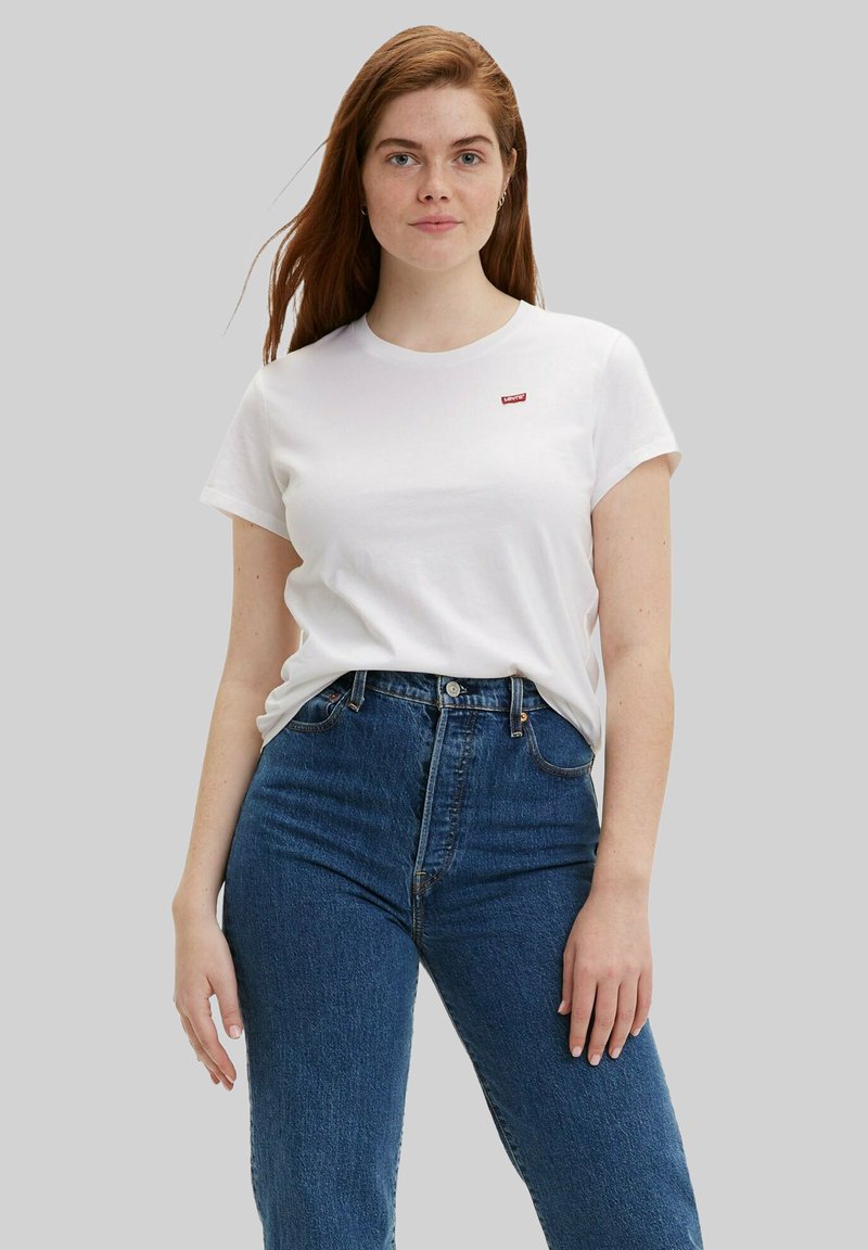 Junge Frau mit roten Haaren, die ein weißes T-Shirt mit kurzen Ärmeln und eine hoch taillierte blaue Jeans trägt, steht vor einem schlichten grauen Hintergrund.