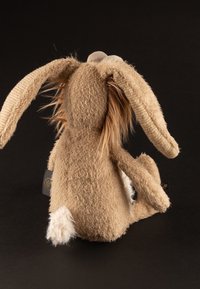 Stuffed rabbit with long, soft tan ears, shaggy fur texture, and white fluffy tail. Tag visible on the side, back view against a dark background.